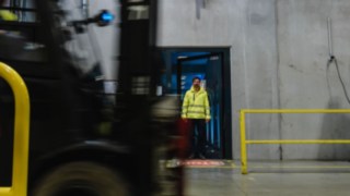 Un homme en tenue de sécurité sort d’une usine par une porte. Un chariot élévateur passe devant lui. Un homme en tenue de sécurité sort d’une usine par une porte. Un chariot élévateur passe devant lui.