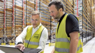 Deux hommes vêtus de gilets de sécurité jaunes se tiennent dans un entrepôt à hauts rayonnages et regardent un ordinateur portable. 