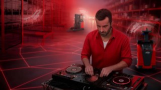 Un DJ se tient devant sa table de mixage dans un entrepôt, avec en arrière-plan des chariots automatisés Fenwick.