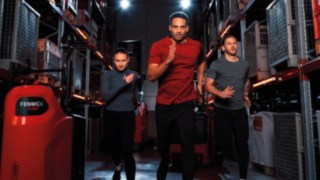 Une femme et deux hommes en tenue de sport courent dans une allée d’entrepôt à côté d'un chariot Fenwick®.