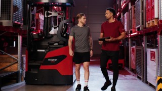 Deux personnes en tenue de sport se tiennent devant un chariot à mât rétractable Ri16 Fenwick   et l’une d’elles tient une tablette à la main.